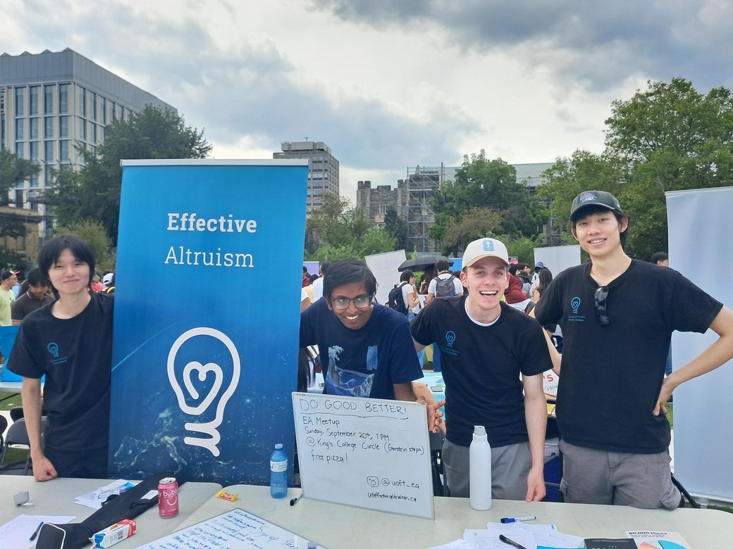 Four UTEA organisers behind the club's table at the U of T clubs fair, beside an Effective Altruism banner and a whiteboard advertising a meetup at King's College Circle.